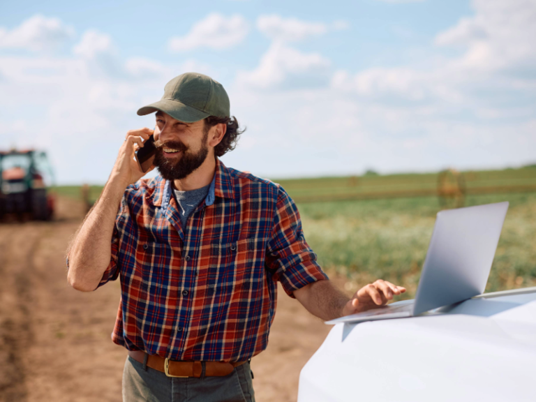 Happy farmer talking on mobile phone while working on laptop in the field.